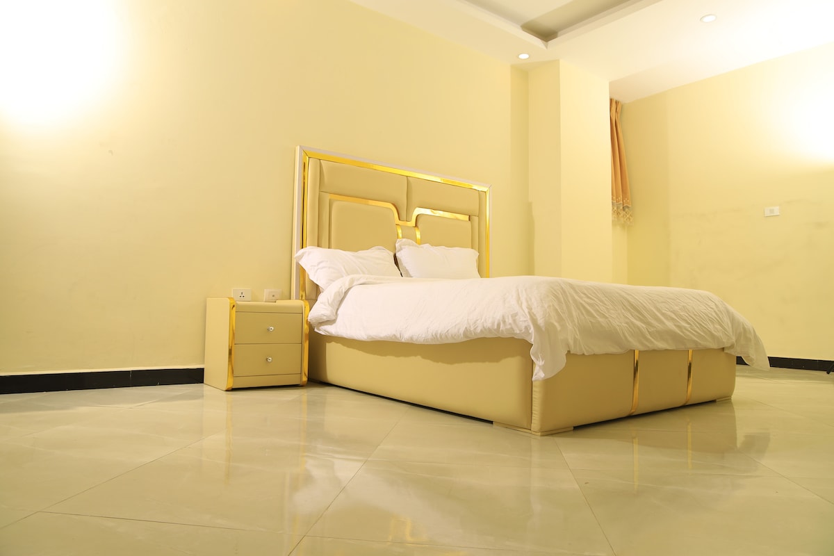A spacious bedroom is featured, showcasing a light-colored bed with a decorative headboard. A bedside table with a simple design is positioned next to the bed. Soft lighting from the walls enhances the warmth of the room, while the floor reflects a polished finish.