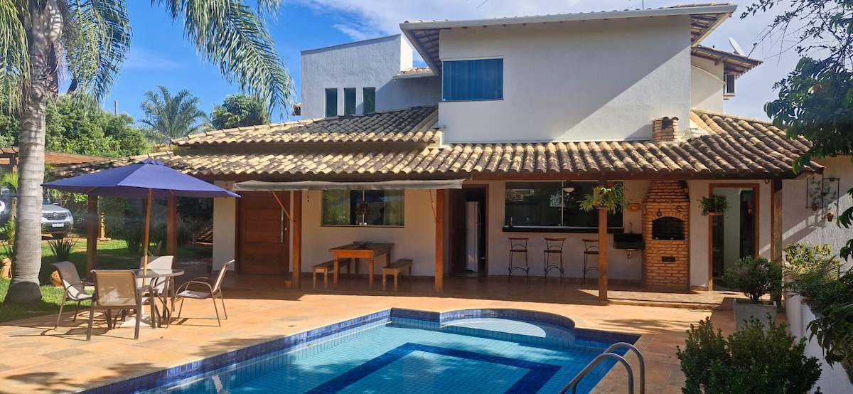 A spacious outdoor area features a swimming pool, surrounded by lounge chairs and a shaded table beneath an umbrella. The patio opens to a large house with a traditional roof and windows, providing a welcoming space for relaxation and enjoyment.