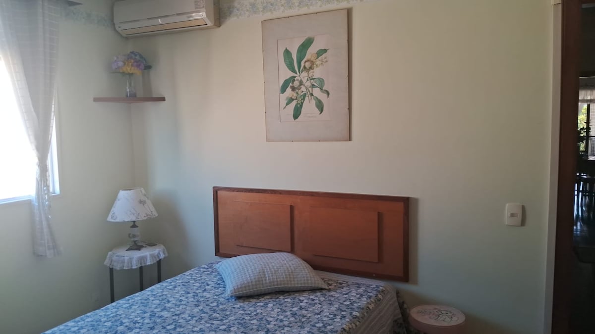 A comfortable guest room is furnished with a spacious bed covered in a patterned blanket. A small bedside table holds a lamp, and a floral wall art piece adds a touch of charm. Natural light enters through a window, enhancing the soft green walls.