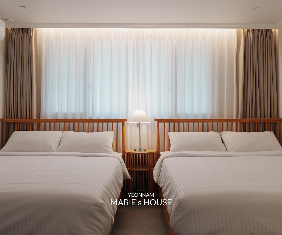 The room features two spacious beds with white linens, set against soft, light-colored walls. Natural light filters through floor-to-ceiling sheer curtains, complemented by a bedside lamp on a wooden nightstand, creating a serene and calming atmosphere.