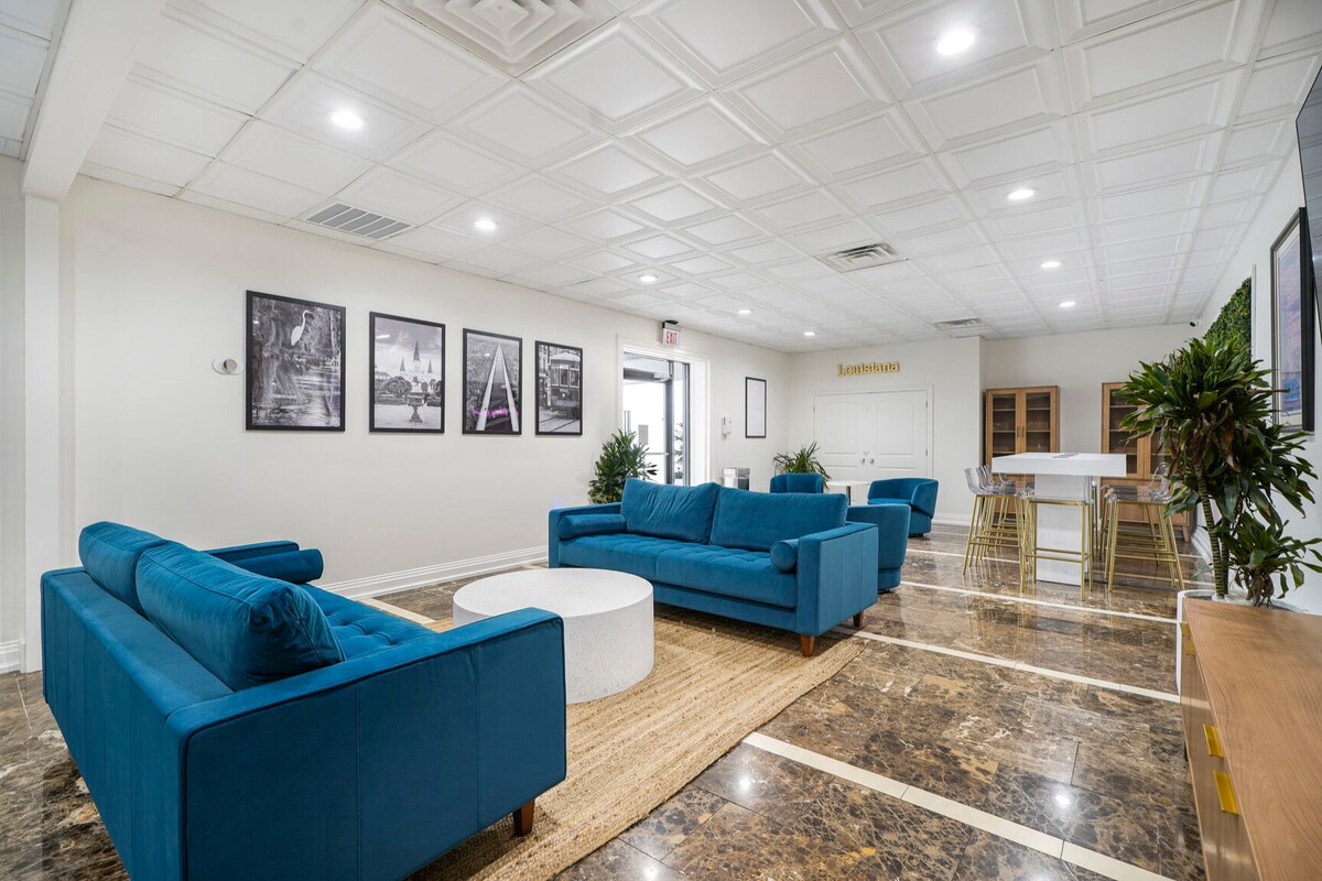 A spacious common area features two blue sofas facing a circular coffee table on a natural fiber rug. Framed black-and-white photographs adorn the walls, while several plants add greenery. A dining table with chairs is located in the background, contributing to a welcoming environment.