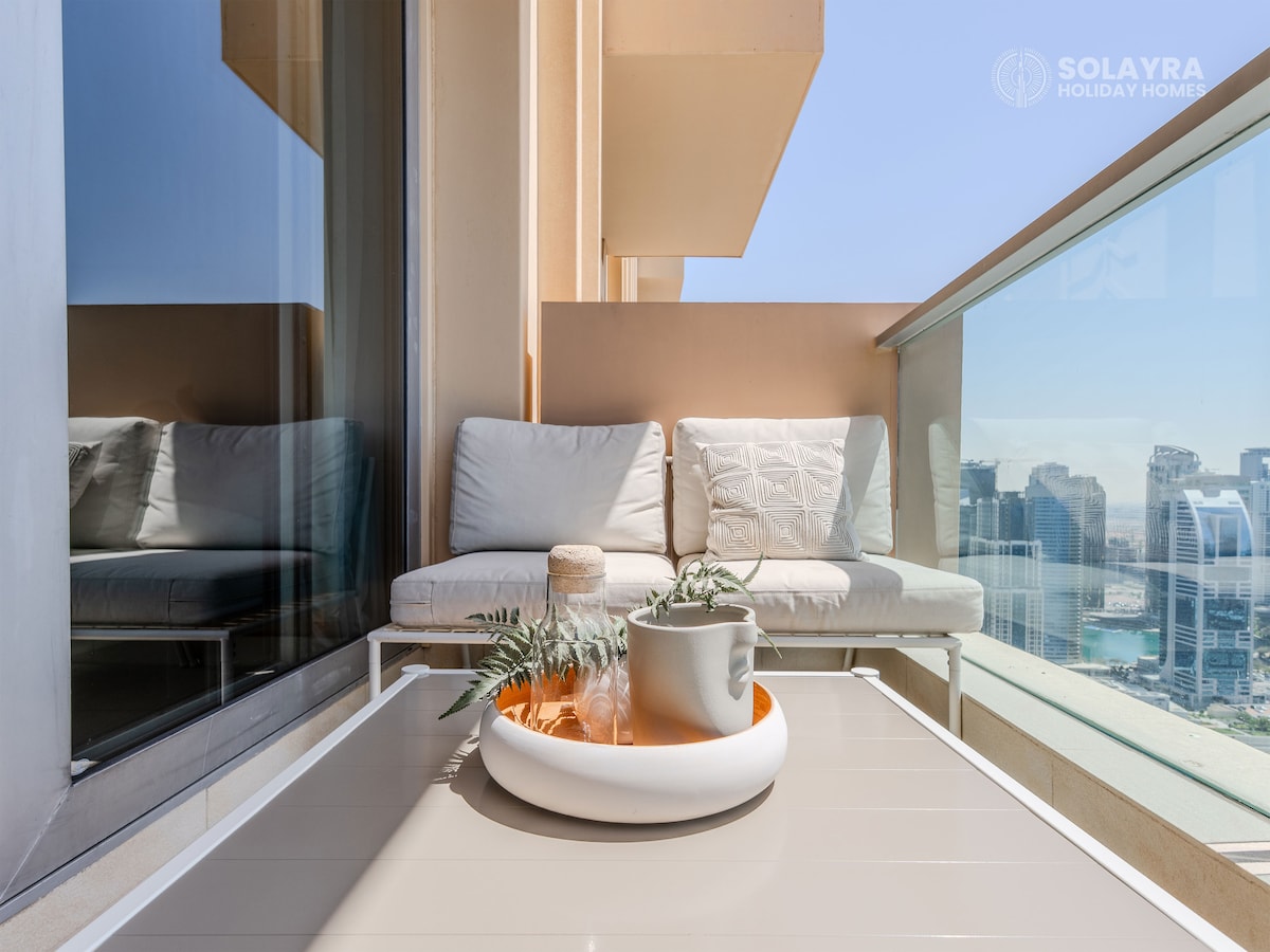 A private balcony features a modern seating area with plush cushions, inviting relaxation. A minimalistic table holds a potted plant and decorative items, all set against a backdrop of stunning city views. Large glass doors enhance the spacious feel and showcase the surrounding skyline.