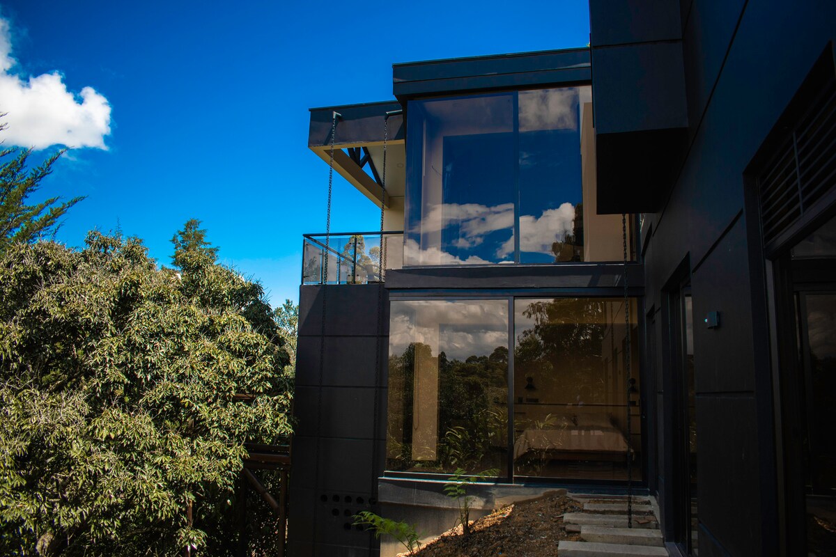 The exterior of a contemporary mountain retreat is highlighted, showcasing large glass windows that reflect the sky and surrounding greenery. A balcony extends outward, emphasizing the connection between indoor and outdoor spaces.