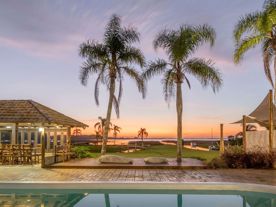 A serene outdoor space features a swimming pool framed by palm trees, with a wooden deck and seating area in the background. The sunset casts a warm glow over the scene, enhancing the tranquil surroundings and offering views of the nearby lagoon.
