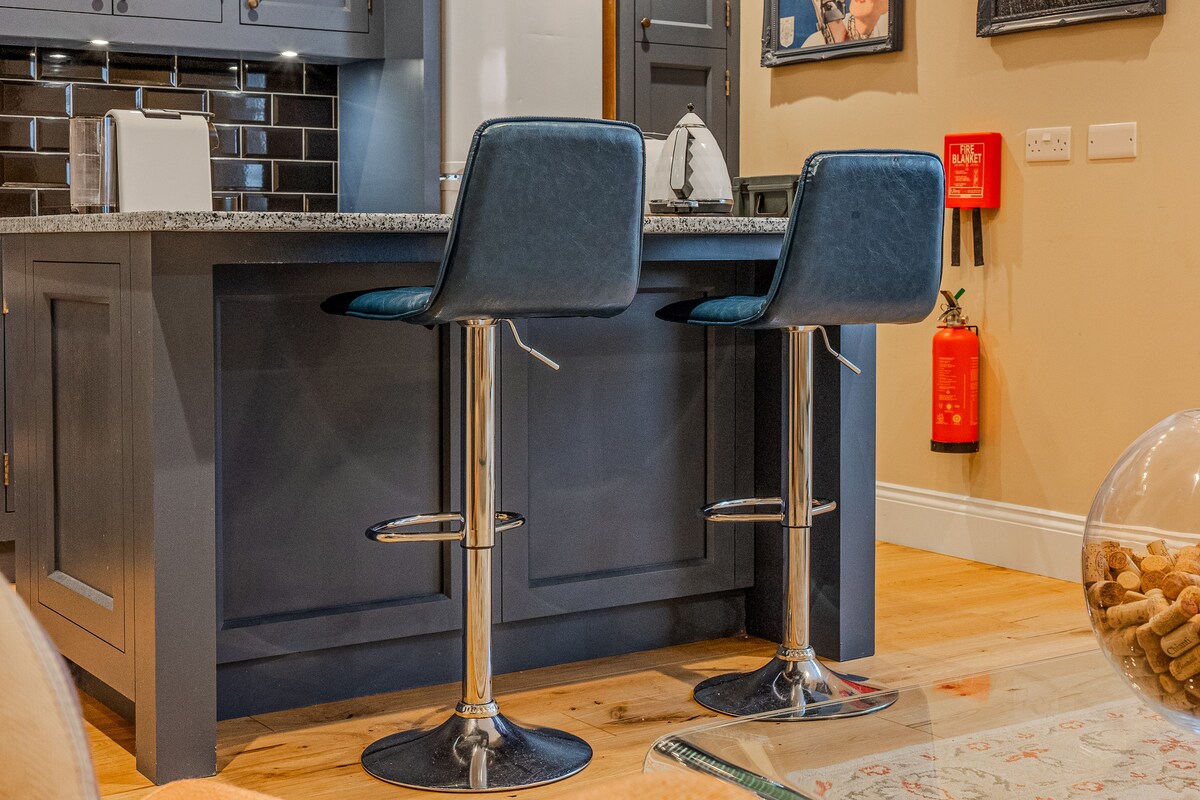 The image features a kitchen island with two sleek bar stools positioned on either side. The island is complemented by a dark countertop, and modern cabinetry can be seen in the background. Safety equipment, including a fire extinguisher, is visible on the wall.