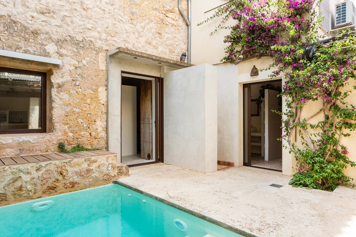 A serene courtyard features a refreshing pool surrounded by natural stone walls. The entrance to a room is visible on the left, and the vibrant greenery of climbing plants adds a touch of color. Light spills onto the textured stone surface, enhancing the tranquil atmosphere.