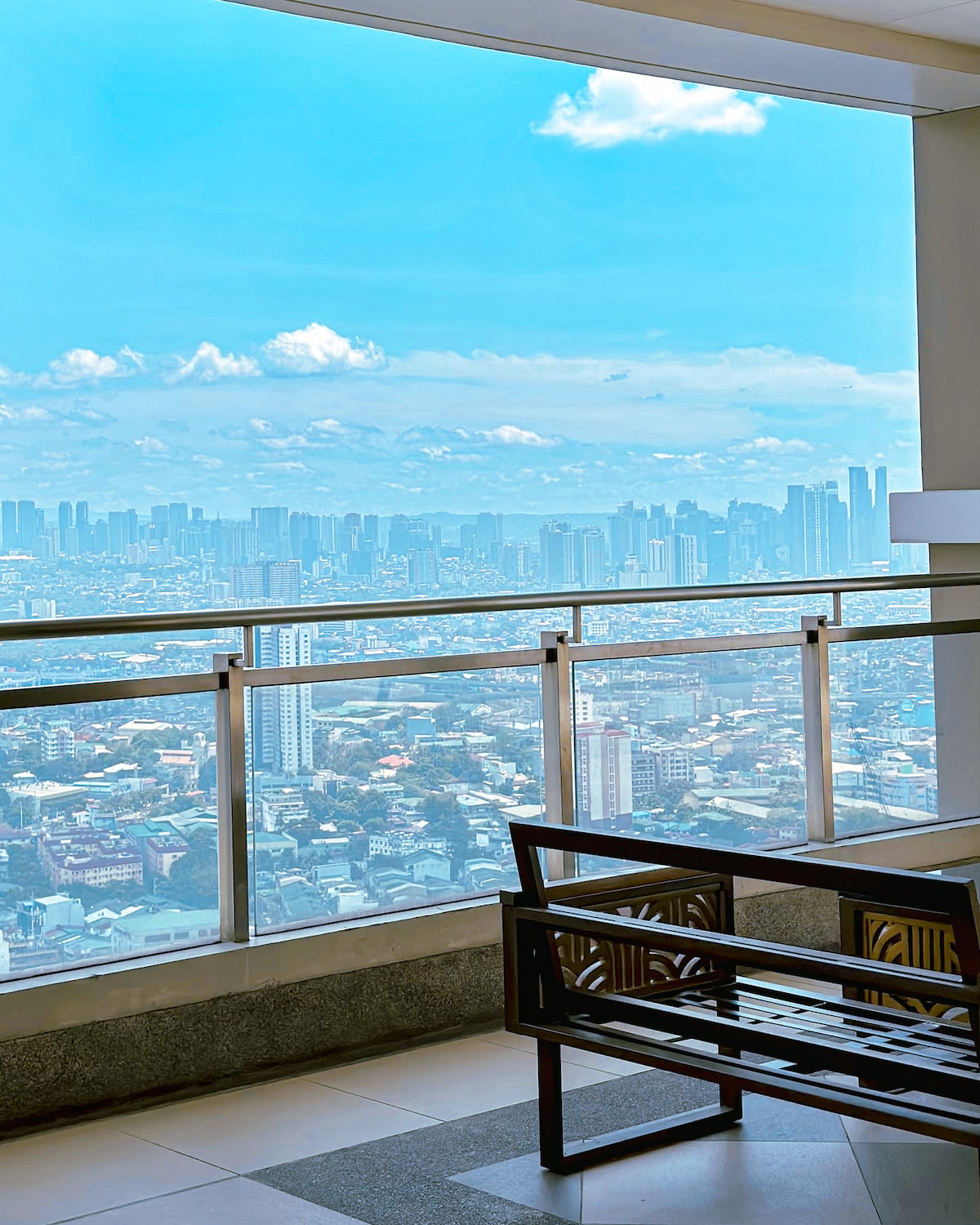 A balcony offers expansive views of the Manila skyline, with a mix of urban buildings and natural landscapes. A metal bench is placed against the glass railing, allowing for a comfortable spot to enjoy the scenery and fresh air.