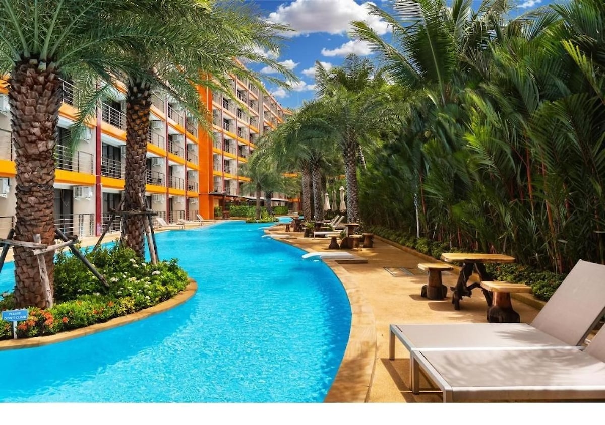 A resort-style pool meanders through lush landscaping, flanked by palm trees and tropical plants. Sun loungers are positioned beside the water, providing a serene space for relaxation. The vibrant orange building appears in the background, creating a lively atmosphere.