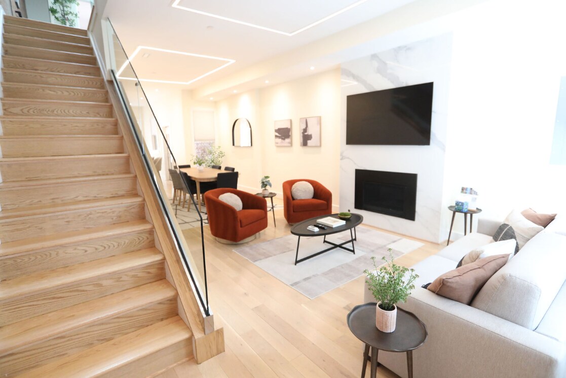 A bright and airy living space features a modern design with wooden flooring and a sleek fireplace. Two cozy armchairs in warm tones are positioned beside a low coffee table. A dining area with a stylish table and chairs is visible in the background, illuminated by natural light.