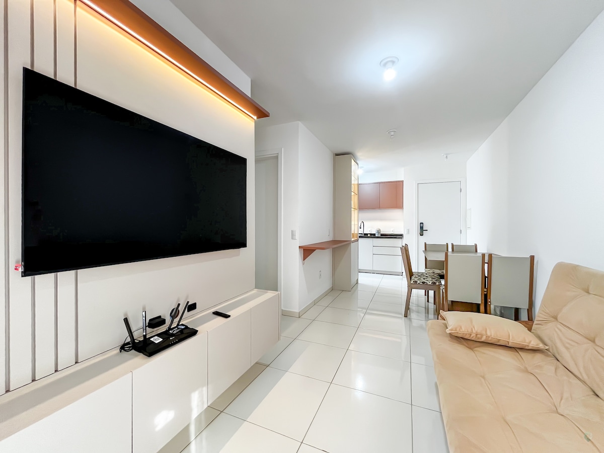 The living area features a large flat-screen TV mounted on a wall with subtle lighting. A comfortable sofa is positioned in front, and a dining table with four chairs is visible in the background. The spacious kitchen is partially seen with sleek cabinetry.