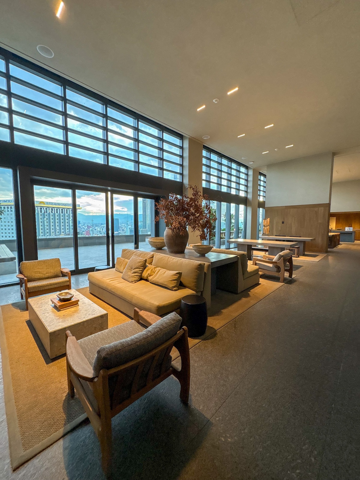 A spacious lounge area is highlighted by large windows, allowing natural light to fill the space. Comfortable seating arrangements, including a sofa and armchairs, surround a central coffee table. Decorative plants add a touch of greenery to the inviting atmosphere.