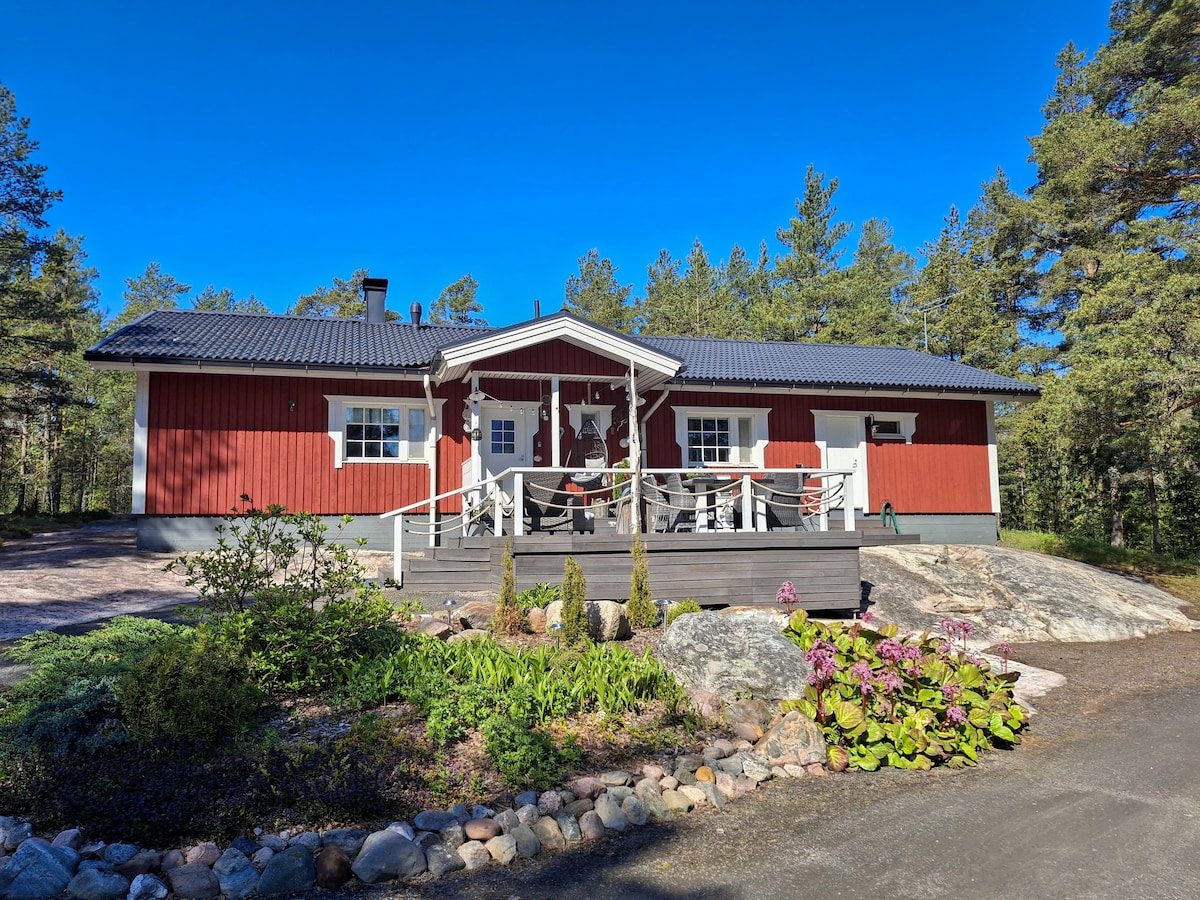 A welcoming red wooden house is set against a backdrop of pine trees. A large deck is accessible from the front, featuring steps leading up to the entrance. It is surrounded by a landscaped area with a variety of plants and natural stone.