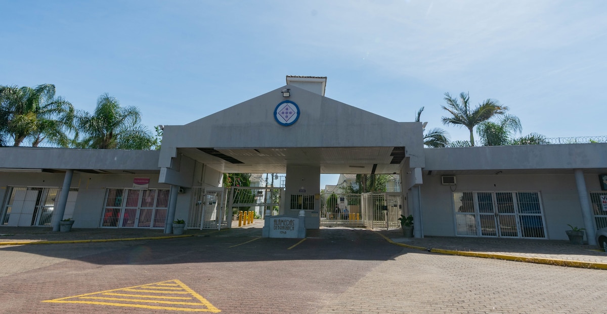 An entrance gate to a condominium complex is shown, featuring a modern architectural style. Palm trees are visible on either side of the pathway leading to the building. A circular logo is mounted above the entrance, and security features, including barriers and a reception area, are evident.