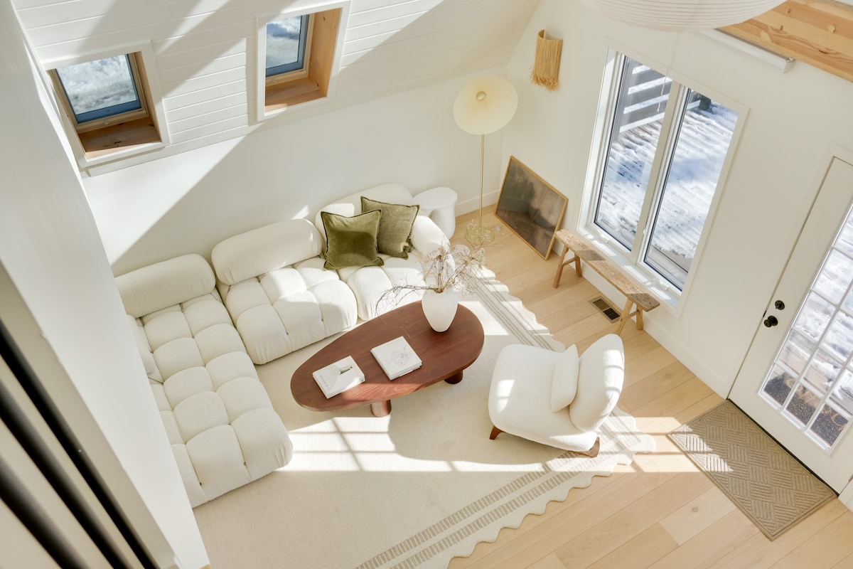 A spacious living area features a creamy sectional sofa paired with a minimalist round coffee table. Natural light streams through large windows, casting soft shadows on the light wooden floor. A cozy armchair is complemented by decorative accents, enhancing a calm and inviting vibe.