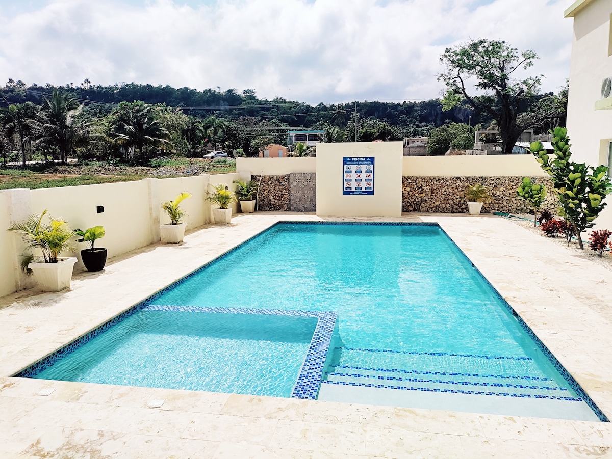 A shared swimming pool features crystal-clear water and a shallow entry with steps. The surroundings are adorned with potted plants, and a tiled wall displays pool rules. Lush greenery is visible in the background, contributing to a serene atmosphere.