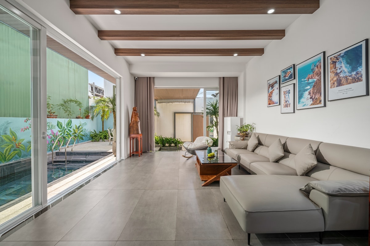 A spacious living area features a light grey sectional sofa paired with a wooden coffee table. Large glass doors open to a private outdoor space, bringing light into the room. Framed artwork decorates the walls, while potted plants add a touch of greenery.
