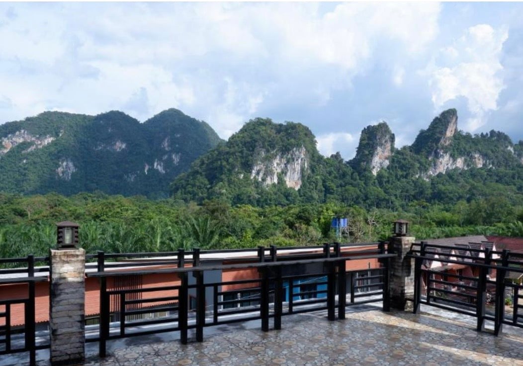 A spacious terrace offers stunning views of lush green mountains, with rocky peaks rising prominently against a cloudy sky. The sturdy railing surrounds the terrace, providing a safe space to admire the natural beauty of the landscape.