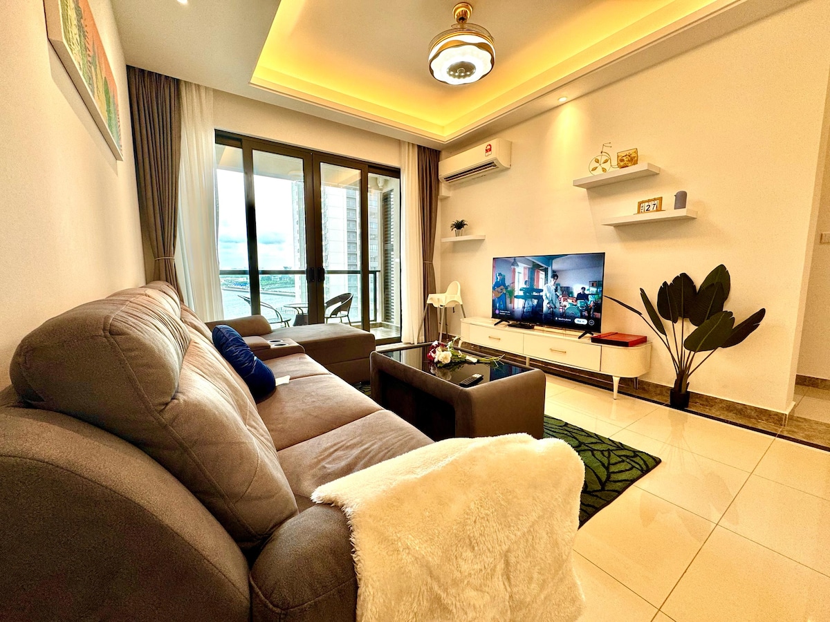 A modern living area features a comfortable gray sofa and a coffee table positioned on a decorative rug. A large television is mounted on the wall, and glass doors open to a balcony, allowing natural light to fill the space.