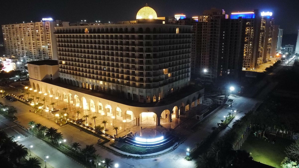 An illuminated building with a distinctive dome stands majestically at night, surrounded by softly glowing lights and palm trees. The architecture features arched columns at the base and multiple floors, with a backdrop of high-rise buildings, creating a captivating urban landscape.