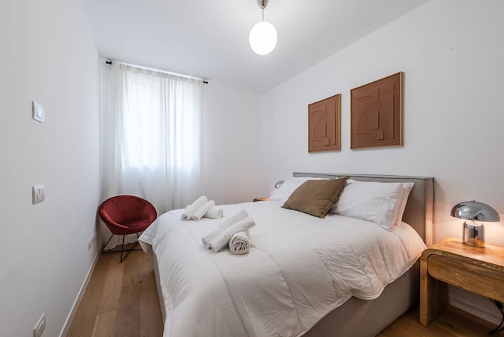 Een intieme en goed ingerichte kamer met een tweepersoonsbed, een matras van 23 cm hoog, houten nachtkastjes, zachte lichten en een bordeauxrode fluwelen stoel voor een stijlvol tintje