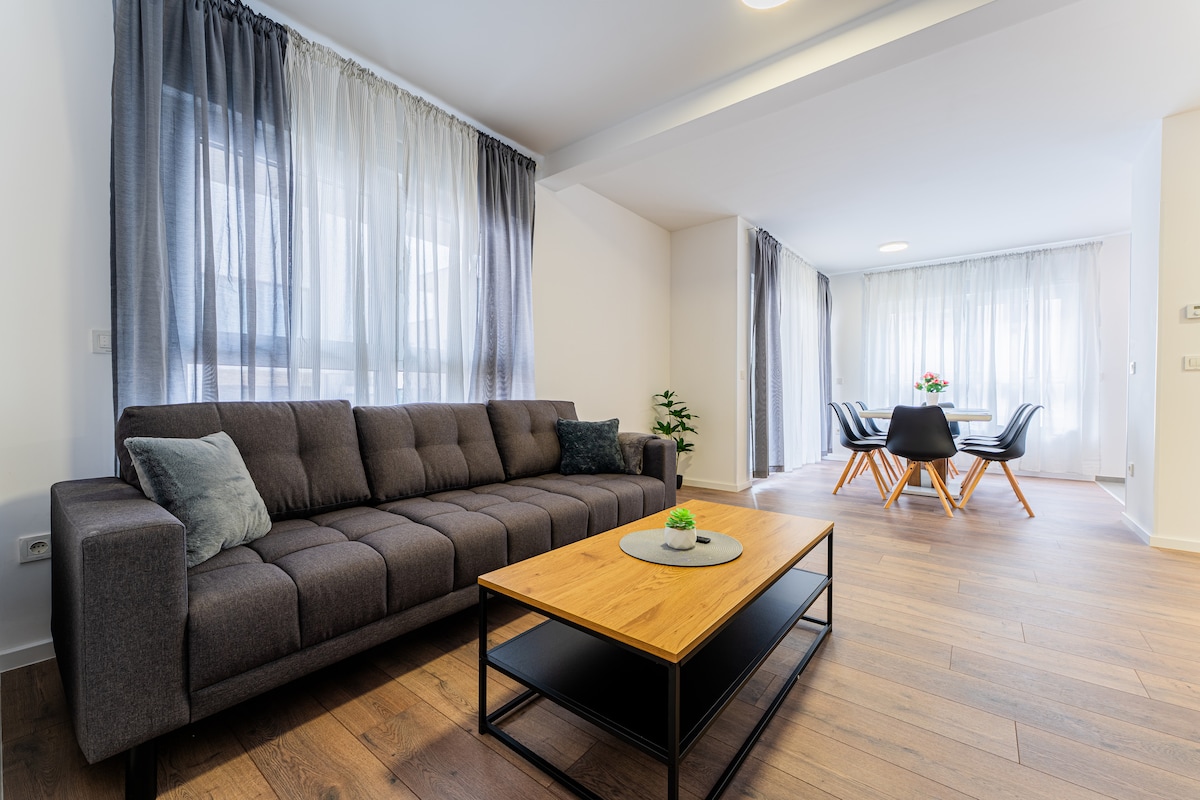 A spacious living area is furnished with a gray sofa and a wooden coffee table, featuring a decorative centerpiece. Large windows with sheer curtains invite natural light, while a dining area with black chairs and a table is visible in the background.