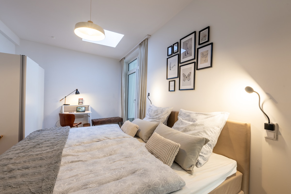 A bright bedroom features a spacious bed adorned with soft bedding and decorative pillows. Framed artwork decorates the wall above the bed. A desk and chair provide a dedicated workspace, while natural light filters in from a skylight and partially covered window.