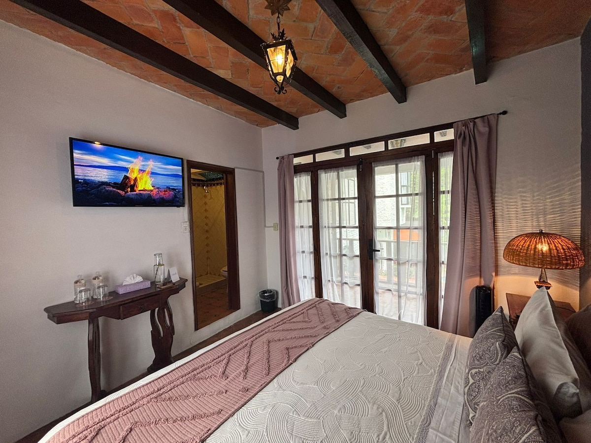 A spacious bedroom is presented, featuring a king-size bed with a neatly arranged blanket. A flat-screen TV is mounted on the wall opposite the bed. Large glass doors provide access to a balcony, while a warm lamp casts a gentle glow on a wooden side table.