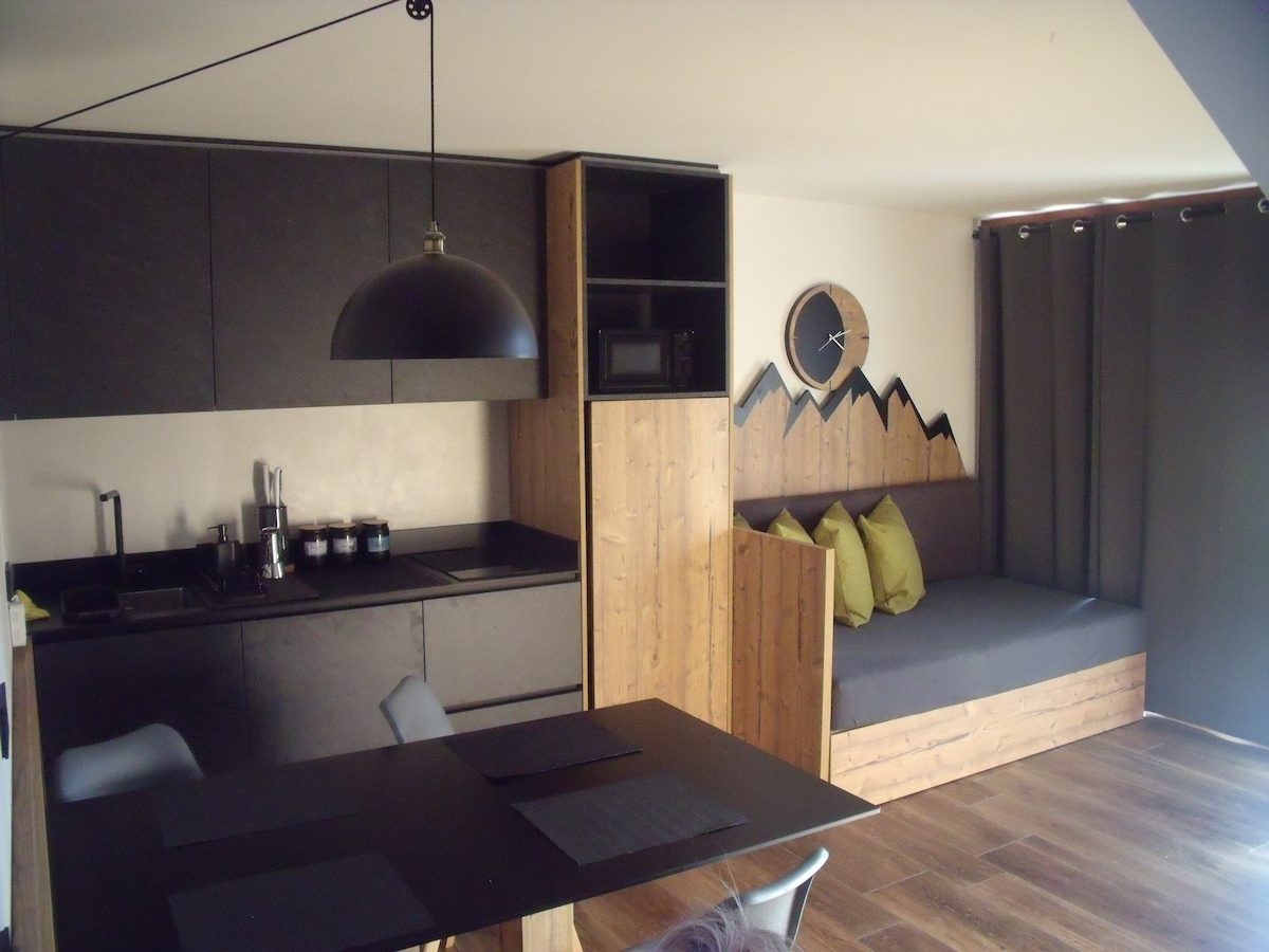 A modern kitchen area includes sleek black cabinetry, a blend of natural wood elements, and a cozy seating nook with green cushions. A minimalist dining table and a round pendant light enhance the space, while a mountain-shaped wooden accent adds a touch of nature indoors.