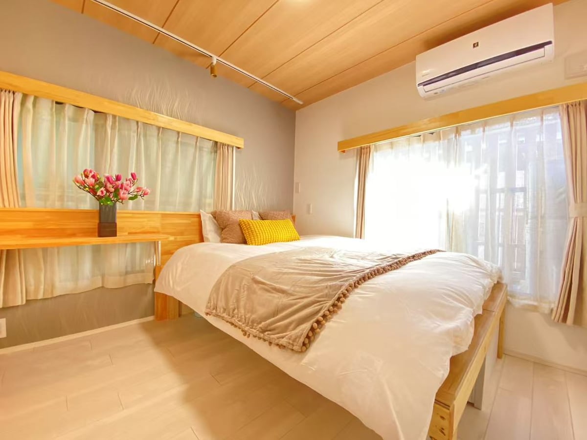 A light-filled bedroom features a spacious bed with soft bedding and decorative pillows. A wooden drawer unit and a vibrant bouquet of flowers add warmth. Natural light streams through sheer curtains, accentuating the room's inviting atmosphere.