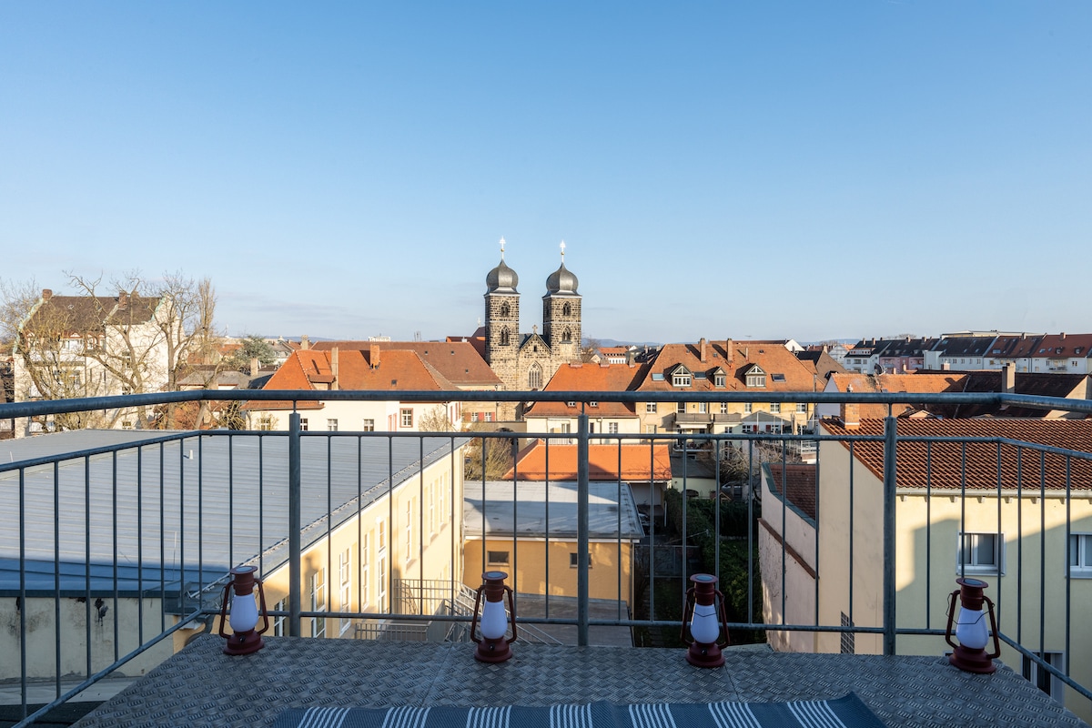 The balcony offers a panoramic view of the surrounding rooftops, showcasing buildings with terracotta roofs. In the distance, two church towers rise prominently against the clear blue sky, providing a glimpse of the local architecture.