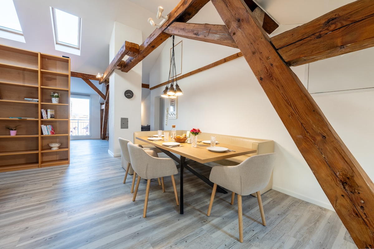 A dining area features a wooden table set for four, complemented by light-colored chairs. Exposed wooden beams create a rustic feel, while natural light floods the space through skylights. A minimalist bookshelf with decorative items is positioned in the background.