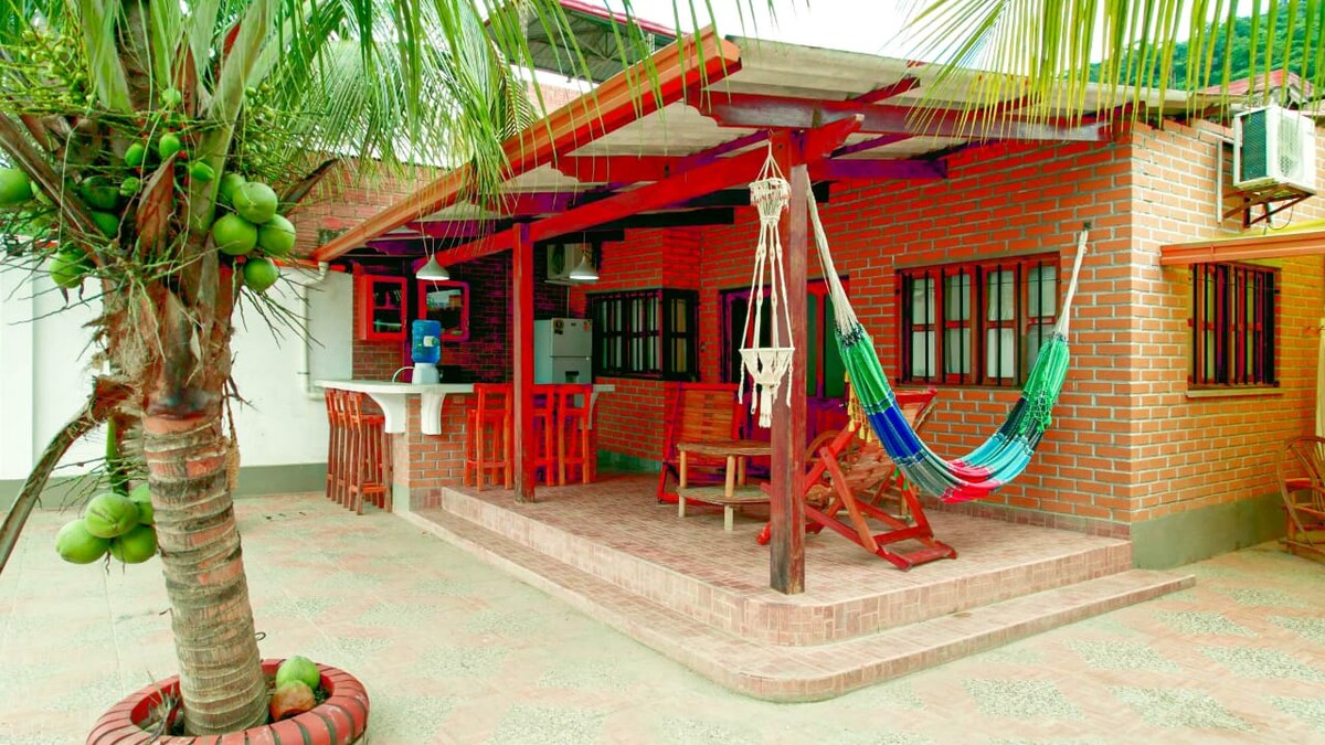 A cozy outdoor seating area features a wooden table and chairs, complemented by a colorful hammock. A palm tree with coconuts stands nearby, and brick walls provide a rustic charm. An air conditioning unit is mounted on the side of the house.