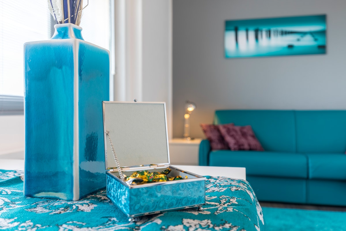 A decorative blue vase stands beside a textured teal tablecloth. An open jewelry box reveals various pieces inside, while a modern sofa with purple cushions is positioned in the background. A vibrant wall artwork adds a touch of color to the neutral-toned walls.