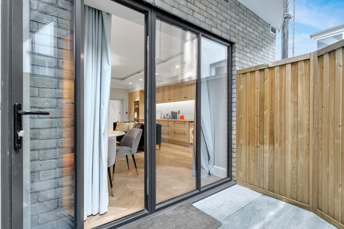 A glass sliding door opens to a private patio area, featuring a light wooden deck and a low fence. Natural light enters from the interior, illuminating the door and the wooden flooring inside. Soft curtains drape gracefully beside the entrance.