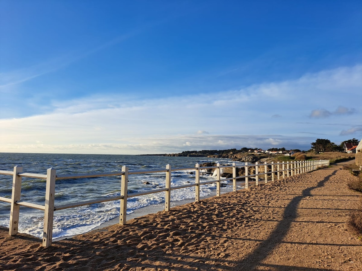 A sandy beach setting is shown with gentle waves lapping at the shore. A white fence runs parallel to the water, offering a clear view of the coast. The sky is bright blue with scattered clouds, contributing to a serene atmosphere.