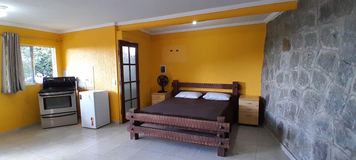A spacious room features a rustic wooden bed with bedding and a small bedside table on each side. A fan and a refrigerator are positioned nearby, while a kitchen area with an oven is visible. Natural light enters through the window, highlighting the textured stone wall.