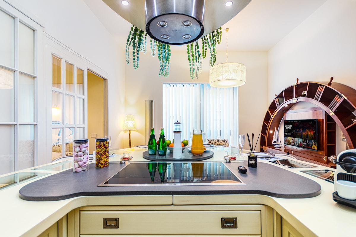 A modern kitchen features a central island with a cooktop, surrounded by decorative items and jars filled with various snacks. Natural light enters through a nearby window, illuminating the area. The space is complemented by a unique shelving unit in the background.