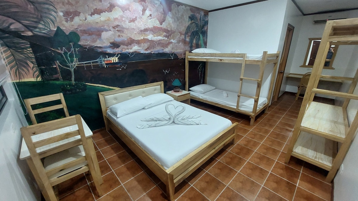 A spacious room features a double bed, a bunk bed, and an additional single bed. Wooden furniture, including a chair and a table, complements the design. A vibrant mural depicting nature adorns one wall, enhancing the overall ambiance. Warm tones from the tiled floor provide a cozy feel.