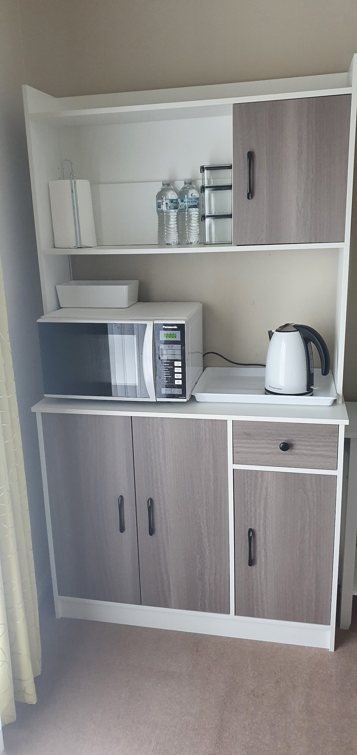 A compact kitchenette features a microwave, electric kettle, and a shelf displaying water bottles. The cabinetry is designed with a modern wood finish and provides ample storage space, contributing to an organized and functional guest area.