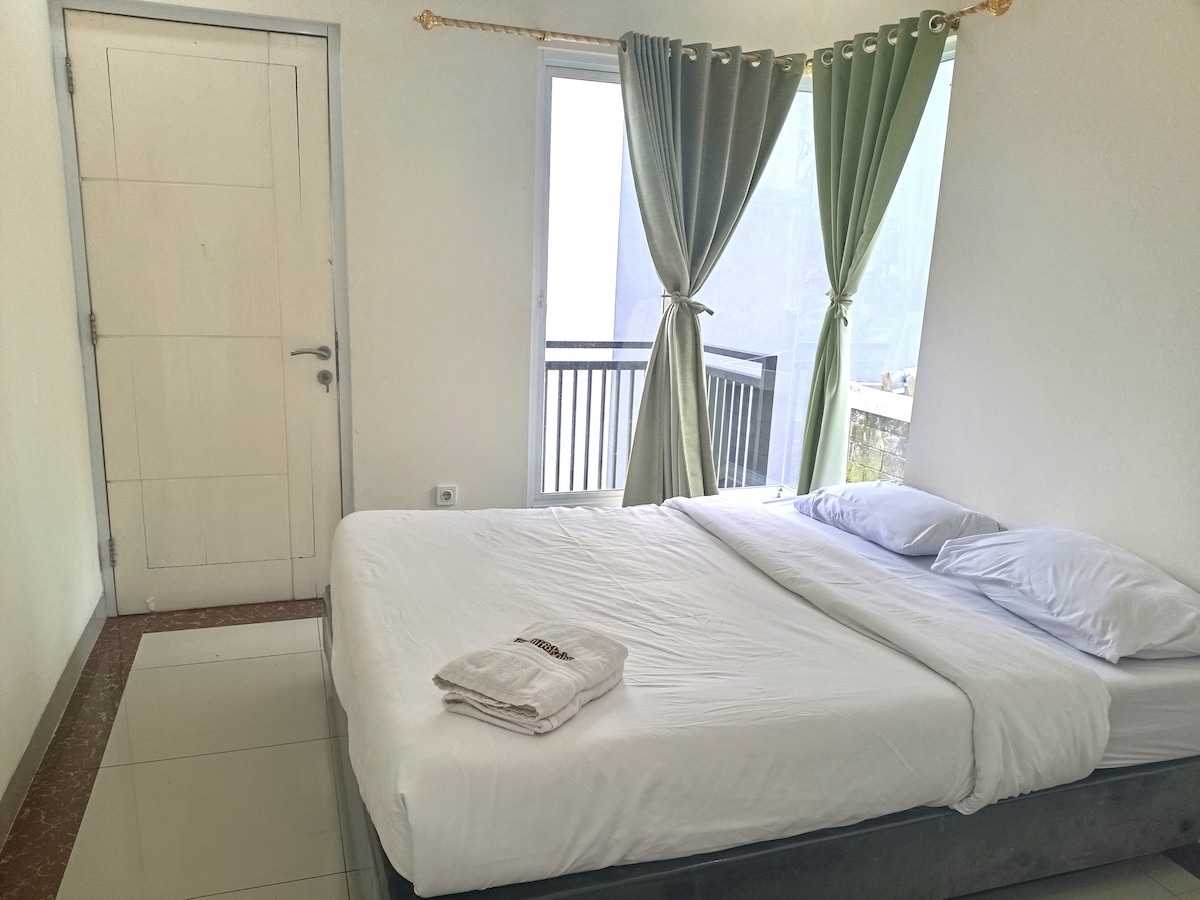 A simple bedroom features a spacious bed topped with crisp white linens. Natural light enters through large windows adorned with green curtains, complementing the neutral walls. A door leads to an outdoor space, enhancing the room's accessibility.