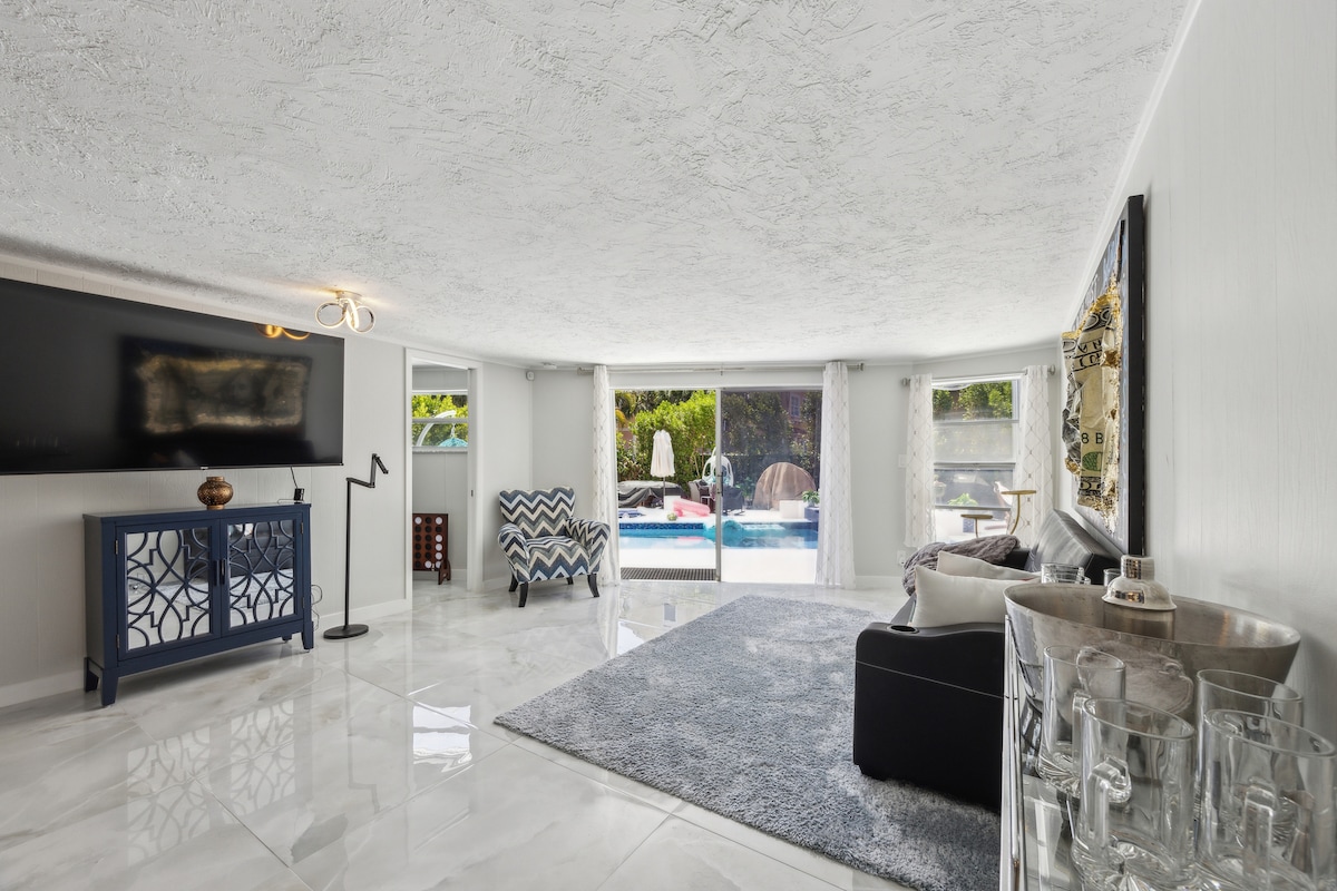 Bright Living Area with Pool View
Spacious and sunlit living room featuring glossy marble floors, a cozy area rug, flat-screen TV, and direct views of the sparkling pool through full-length sliding doors.