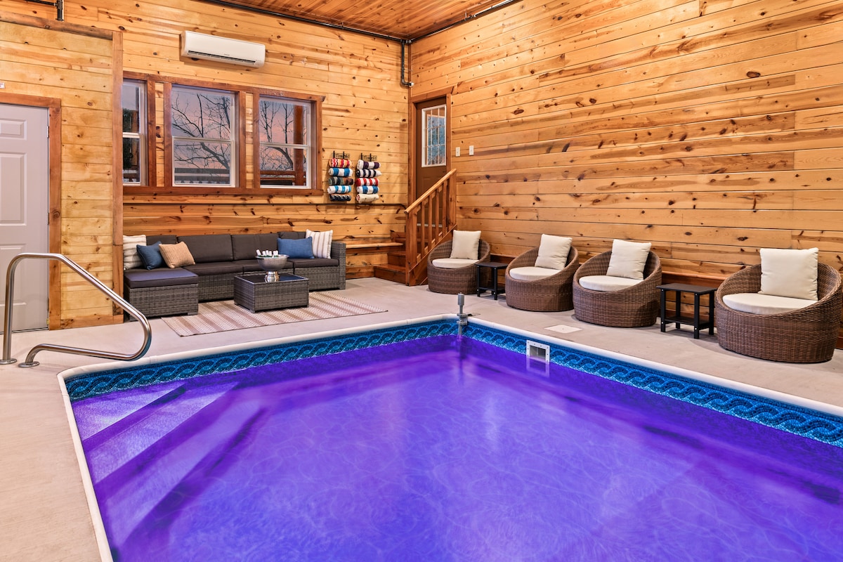 An indoor heated swimming pool is featured, surrounded by wooden paneling which enhances the rustic ambiance. Comfortable seating is available nearby, consisting of a sectional sofa and individual chairs. Light filters through large windows, offering views of the outdoors while adding a warm glow to the space.