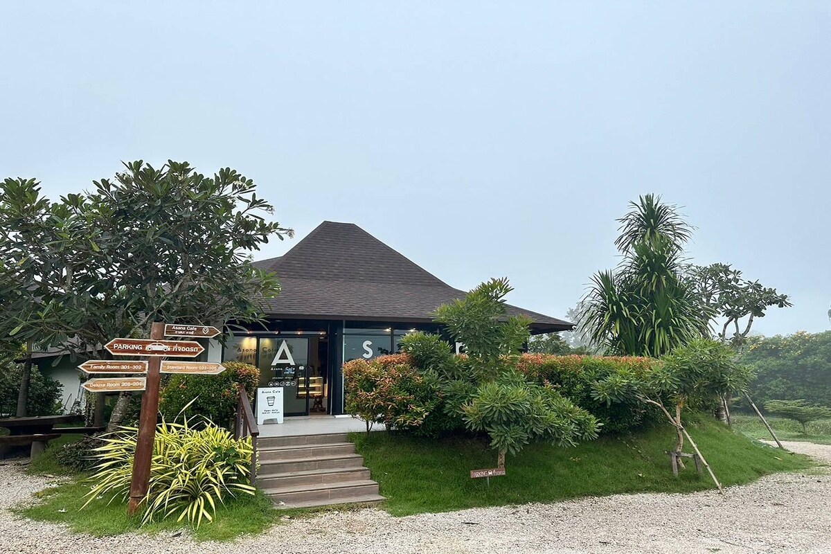 A modern building with a distinctive rounded roof is surrounded by lush greenery and well-maintained plants. A set of stair steps leads to the entrance, while directional signs provide guidance to various locations. The atmosphere is enhanced by a tranquil, misty environment.