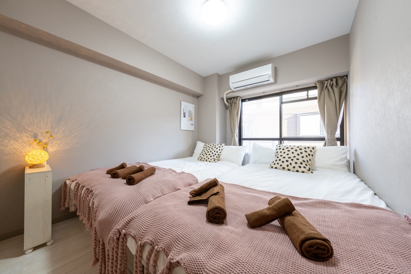 A well-lit bedroom features two semi-double beds with soft pink throws and neatly arranged brown towels on each. Plush pillows with patterned covers are positioned on the beds. Natural light filters in through the window, complemented by soft beige curtains.