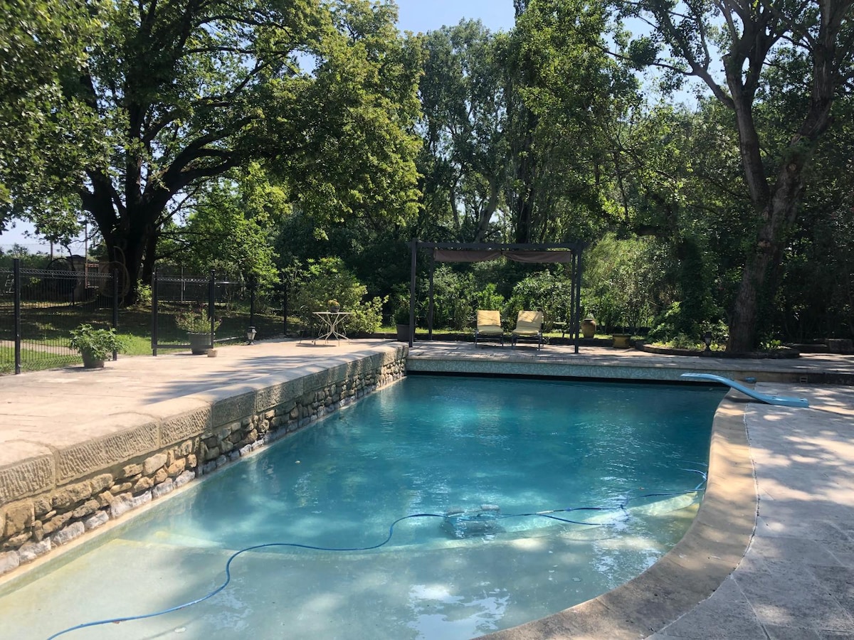 The outdoor pool is surrounded by a spacious terrace, featuring stone accents. Lush greenery and trees frame the area, offering shade and a natural setting. A covered seating area is visible, providing a relaxing spot for outdoor enjoyment.