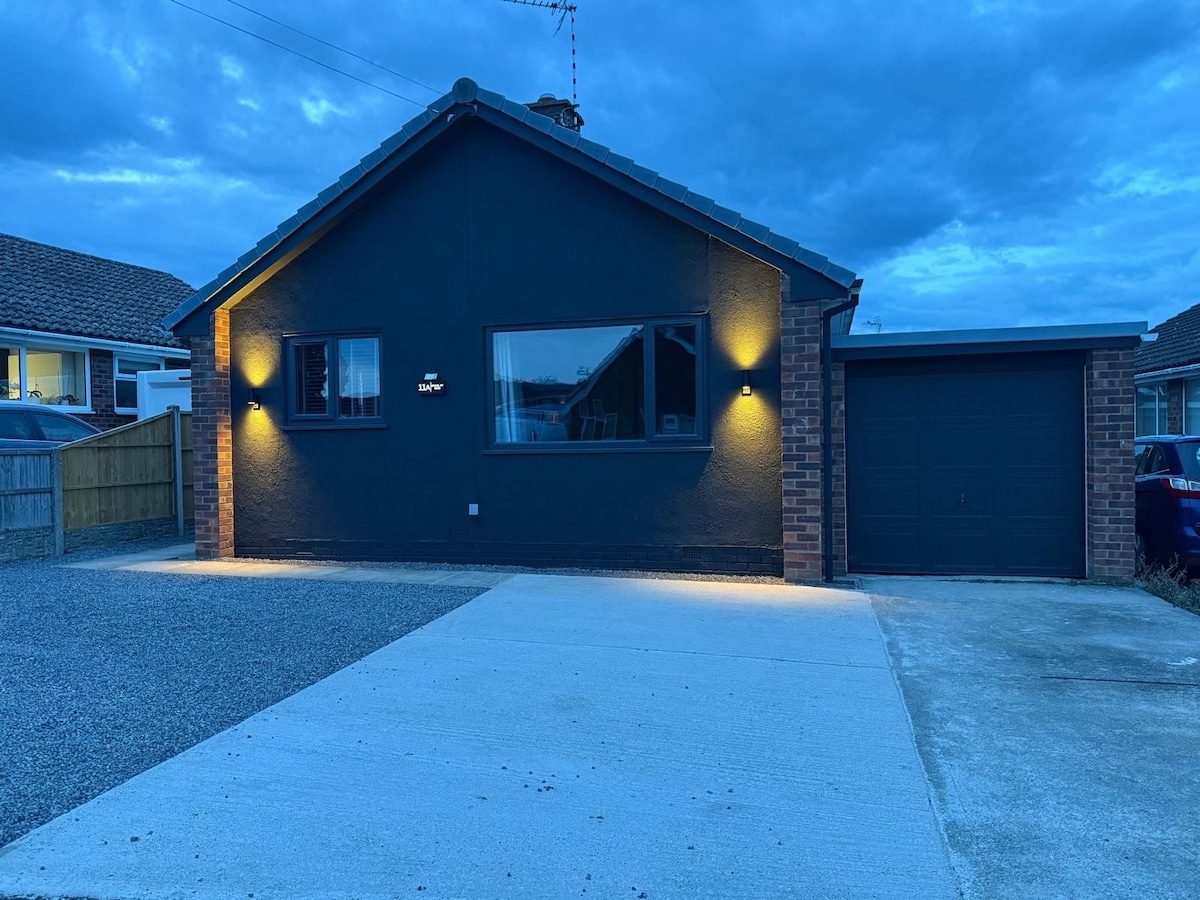 The exterior of the dwelling is visible, showcasing a single-story structure with a dark exterior and a prominent large window. Soft lighting highlights the entrance area. A spacious driveway leads up to a garage, with gravel and concrete surfaces contributing to the welcoming approach.