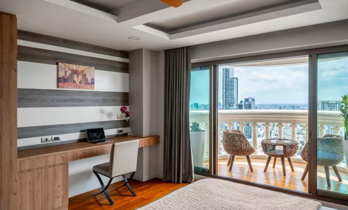 A bright room is presented, featuring a streamlined workspace with a desk and chair. Large windows frame a view of the cityscape, while a small balcony with two cozy chairs overlooks the surroundings. Subtle decor adds a touch of elegance to the space.