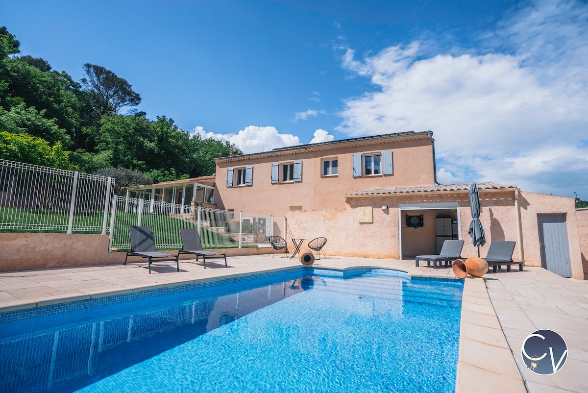 A private pool is surrounded by a spacious sun deck, featuring several lounge chairs. The villa is visible in the background, showcasing a two-story structure with a beige façade and windows. Lush greenery is set against a clear blue sky.