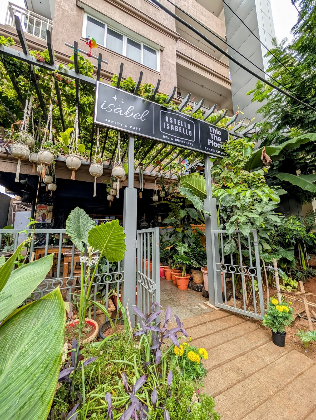 The entrance to Ostello Isabello is surrounded by lush greenery and potted plants. A welcoming gate opens to a quaint patio area, featuring signage for Isabel Bakery & Café and This The Place. The space is infused with natural light, hinting at a cozy atmosphere.