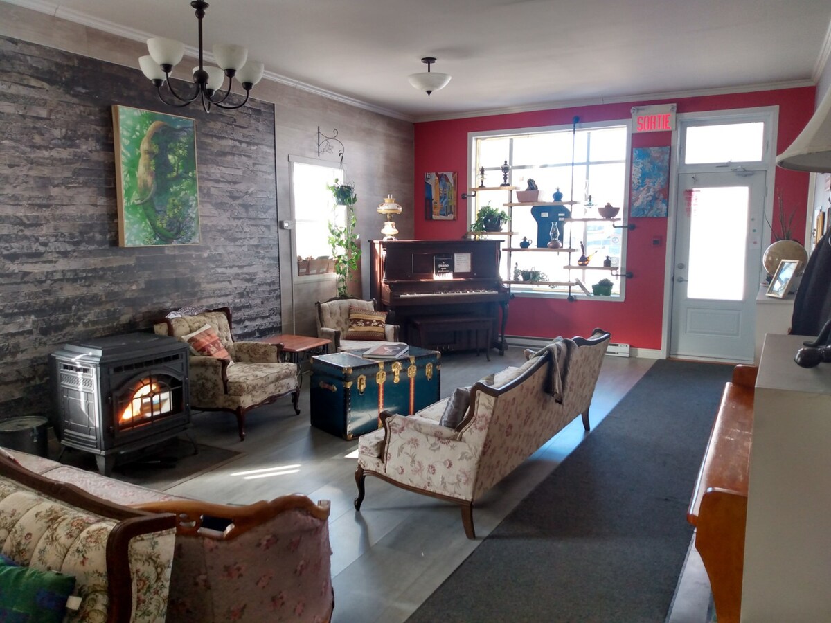 A welcoming common area features vintage furniture, including floral-patterned chairs and a sofa, complemented by a decorative chest and a piano. A warm fire is seen in a cast-iron stove, with plants and artwork adding character to the vibrant red and textured wall.